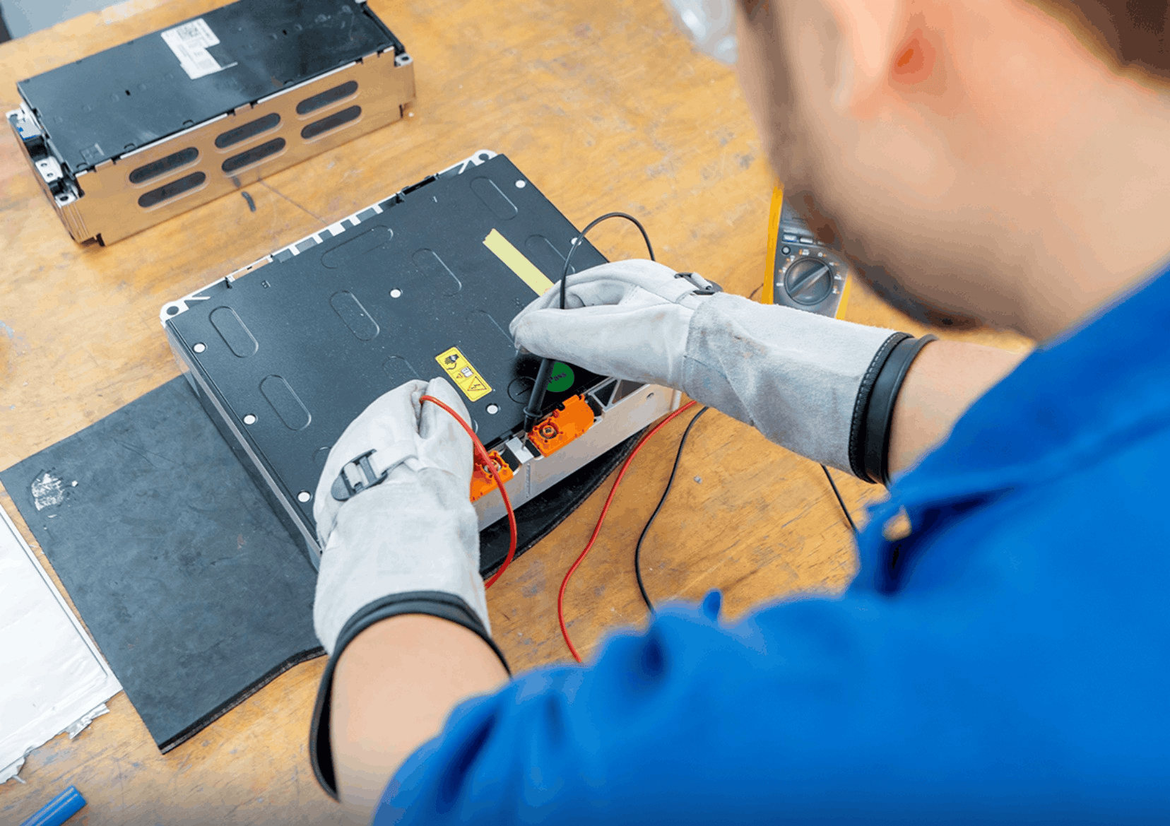 Profissional com luvas de proteção utiliza as pontas de prova de um multímetro para realizar medições elétricas em uma bateria sobre uma bancada de trabalho.